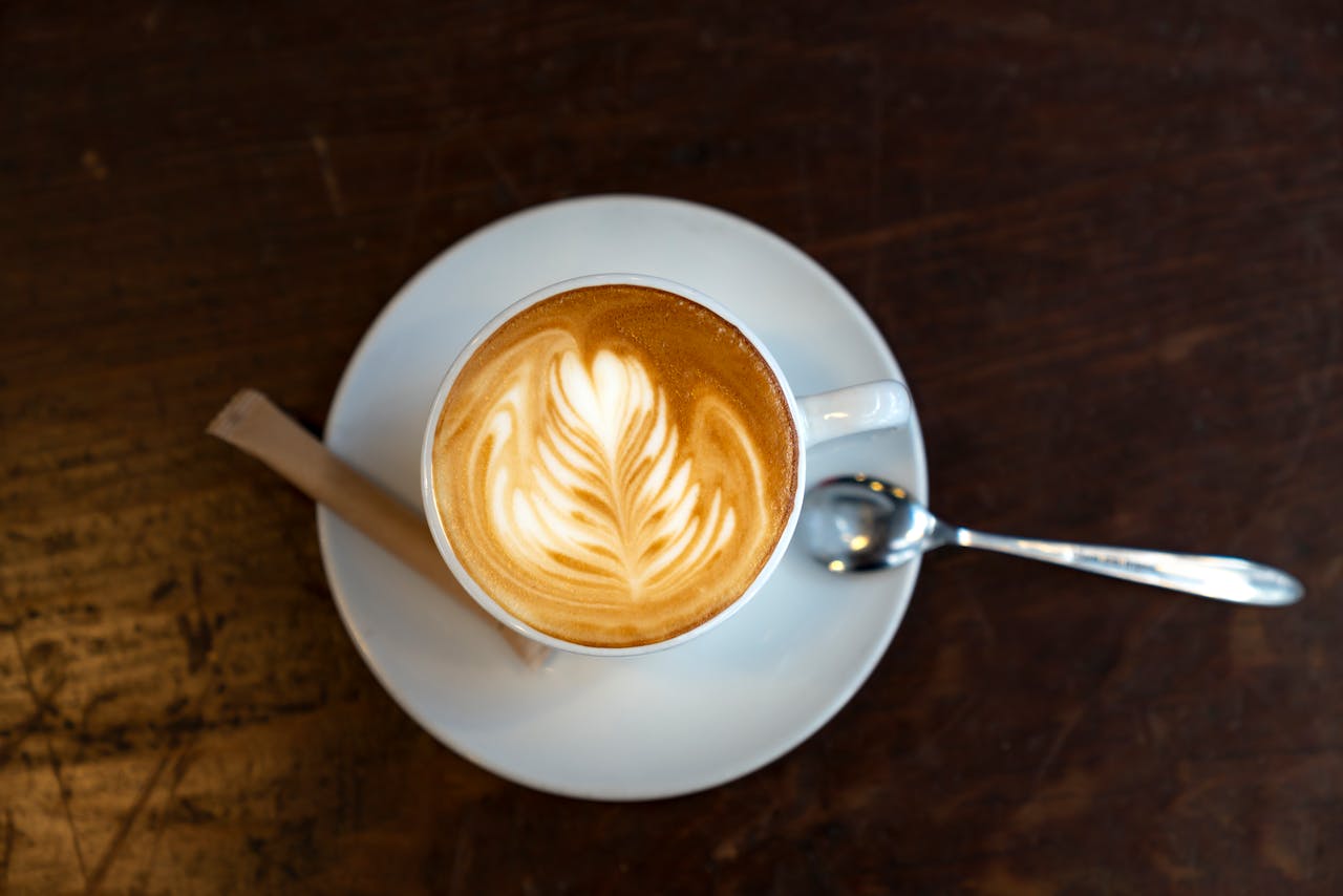 Top-down image of a cappuccino with latte art on a wooden table, served with spoon and sugar.